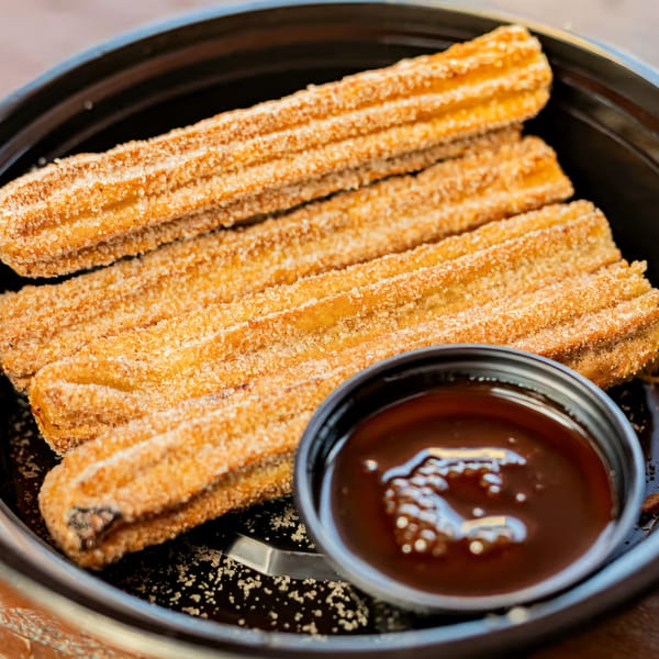 Golden-brown churros, coated in sugar, arranged in a black bowl with a side of chocolate dipping sauce.