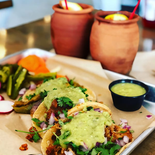 Three La Penca Azul tacos with guacamole, cilantro, and onions, served with pickled vegetables and drinks.