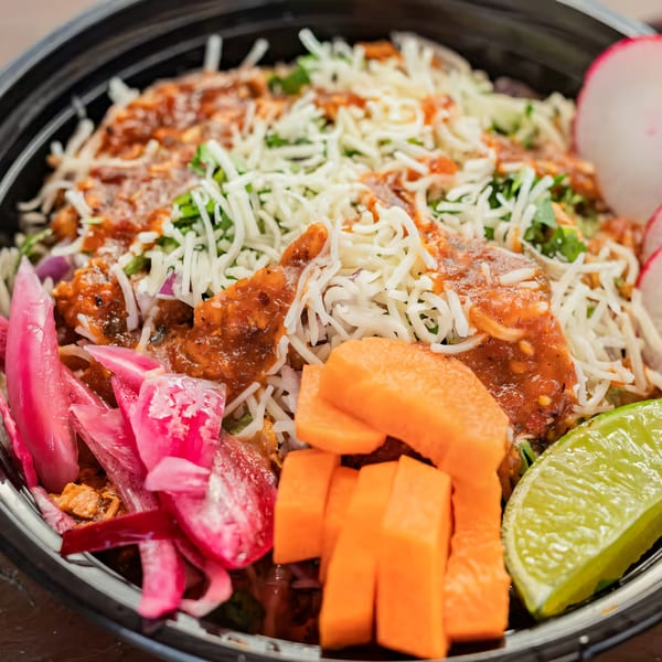 Mexican burrito bowl with shredded cheese, red sauce, pickled onions, papaya, and a lime wedge.