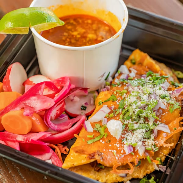 La Penca Azul birria tacos with consommé, pickled radishes, and carrots in a black takeout container.